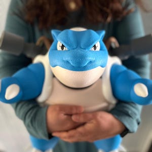 May include: A blue and white Blastoise figure with a detailed, textured design. The figure has two large cannons on its back and is held in front of a person wearing a green shirt. The figure's eyes are white with black pupils.
