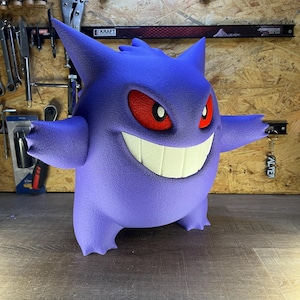 May include: A 3D-printed purple Gengar figure with red eyes and a wide, toothy grin. The figure has outstretched arms and is standing on a wooden surface. The background includes tools and a sign that says "KRAFT".