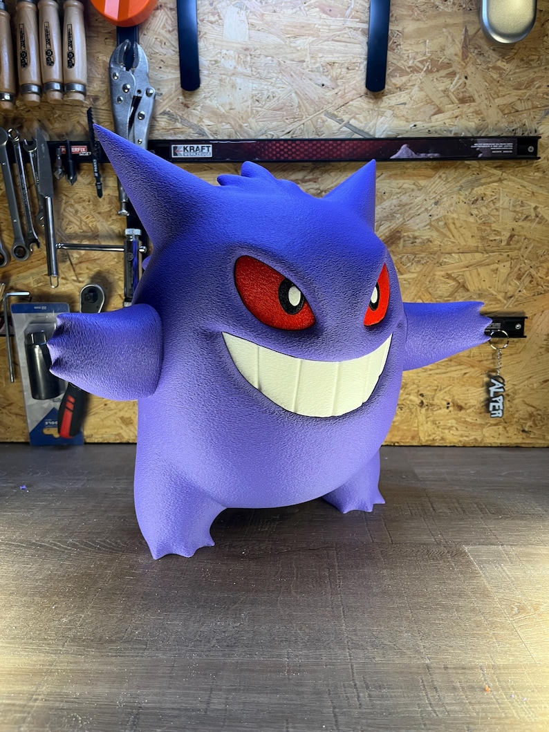 May include: A 3D-printed purple Gengar figure with red eyes and a wide, toothy grin. The figure has outstretched arms and is standing on a wooden surface. The background includes tools and a sign that says "KRAFT".