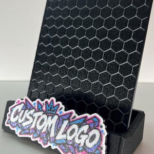 K&ouml;nnte beinhalten: Ein schwarzer Pr&auml;sentationsst&auml;nder mit einem strukturierten Wabengittermuster auf der R&uuml;ckwand. An der Vorderseite befindet sich ein Schild mit dem Text CUSTOM LOGO in Wei&szlig;, umrandet von einem glitzernden blauen, rosa und violetten Graffiti-Design.