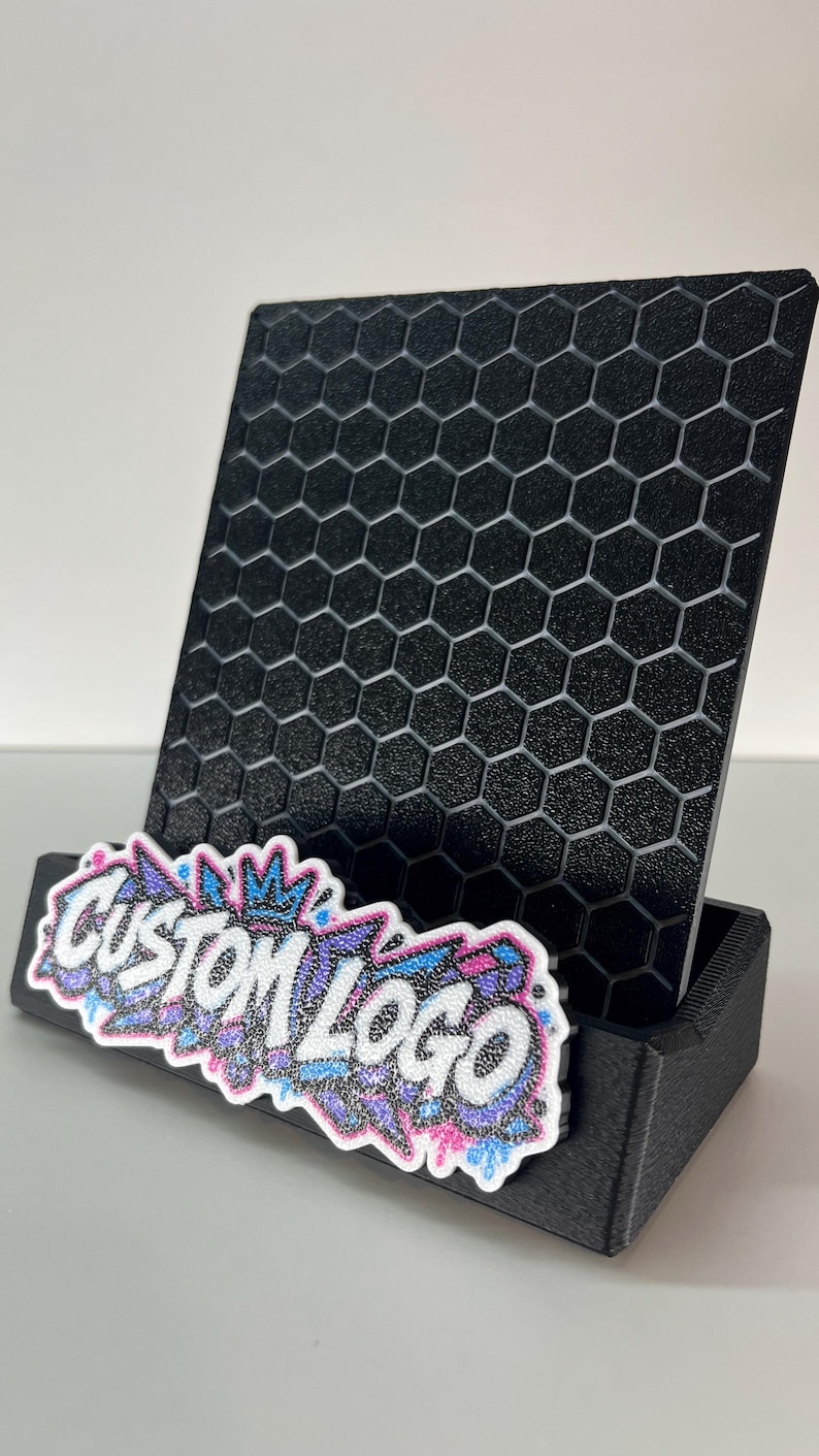 K&ouml;nnte beinhalten: Ein schwarzer Pr&auml;sentationsst&auml;nder mit einem strukturierten Wabengittermuster auf der R&uuml;ckwand. An der Vorderseite befindet sich ein Schild mit dem Text CUSTOM LOGO in Wei&szlig;, umrandet von einem glitzernden blauen, rosa und violetten Graffiti-Design.