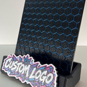 K&ouml;nnte beinhalten: Ein schwarzer Pr&auml;sentationsst&auml;nder mit texturiertem Sechseckmuster und blauen Akzenten. Die vordere Basis zeigt ein wei&szlig;es, rosa und blaues Graffiti-Schild mit dem Text CUSTOM LOGO in einer fetten, stilisierten Schrift mit Glitzereffekt.