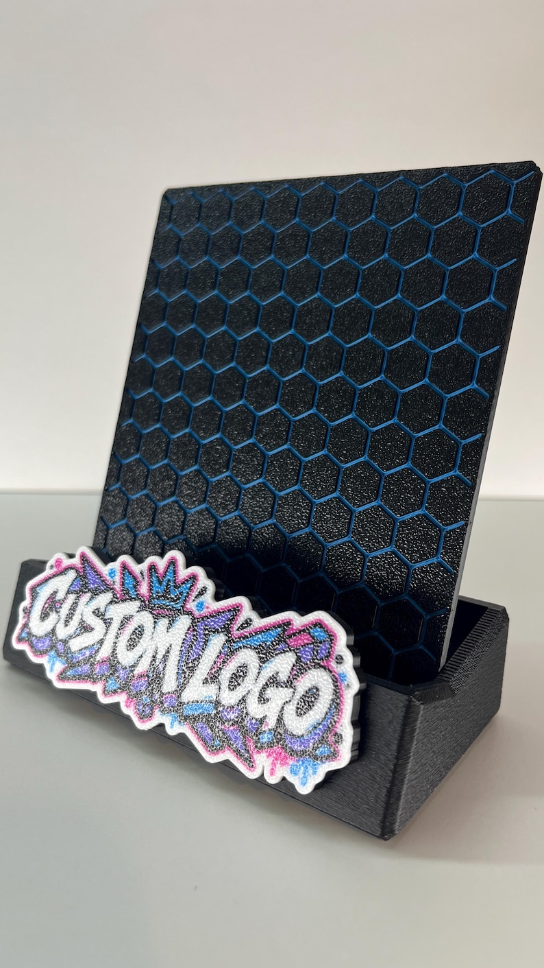 K&ouml;nnte beinhalten: Ein schwarzer Pr&auml;sentationsst&auml;nder mit texturiertem Sechseckmuster und blauen Akzenten. Die vordere Basis zeigt ein wei&szlig;es, rosa und blaues Graffiti-Schild mit dem Text CUSTOM LOGO in einer fetten, stilisierten Schrift mit Glitzereffekt.