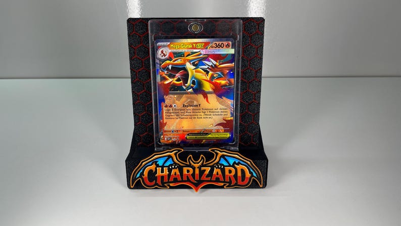 May include: A Mega Charizard Y trading card encased in a clear magnetic protective holder, displayed on a black stand with a red honeycomb pattern and the word CHARIZARD in stylized orange and blue lettering at the base.