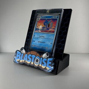 Puede incluir: Un soporte de exhibici&oacute;n negro con patr&oacute;n hexagonal que sostiene una carta coleccionable de Wartortle en una funda protectora transparente. El frente del soporte muestra la palabra BLASTOISE en letras azules, blancas y marrones con dos accesorios tipo ca&ntilde;&oacute;n.