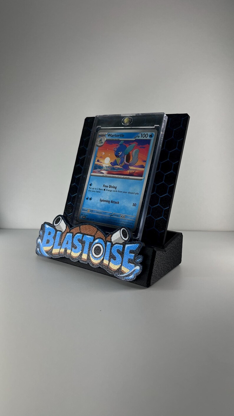 Puede incluir: Un soporte de exhibici&oacute;n negro con patr&oacute;n hexagonal que sostiene una carta coleccionable de Wartortle en una funda protectora transparente. El frente del soporte muestra la palabra BLASTOISE en letras azules, blancas y marrones con dos accesorios tipo ca&ntilde;&oacute;n.