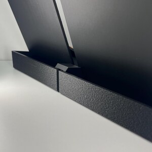May include: A black display stand featuring two upright rectangular panels with smooth surfaces, held in place by a textured, low-profile base. The structure is composed of matte black material, providing a stable support for items.