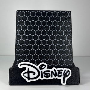 May include: A black display stand featuring a textured honeycomb pattern on the back panel. The front base of the stand is adorned with the white Disney logo in its signature script font, providing a clear contrast against the black material.