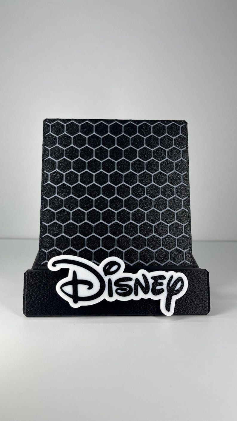 May include: A black display stand featuring a textured honeycomb pattern on the back panel. The front base of the stand is adorned with the white Disney logo in its signature script font, providing a clear contrast against the black material.
