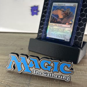 Op de afbeelding: Een Magic The Gathering logo bord in blauw, wit en zwart staat voor een zwarte displaystandaard met een ruilkaart in een beschermhoes. De achtergrond bevat diverse kleurrijke stickers, waaronder Gengar, Venusaur en Pokemon-logo's.