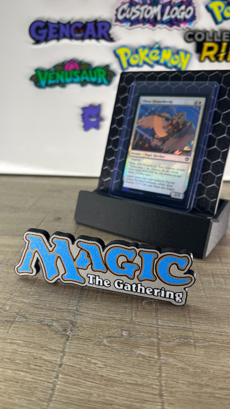 Op de afbeelding: Een Magic The Gathering logo bord in blauw, wit en zwart staat voor een zwarte displaystandaard met een ruilkaart in een beschermhoes. De achtergrond bevat diverse kleurrijke stickers, waaronder Gengar, Venusaur en Pokemon-logo's.