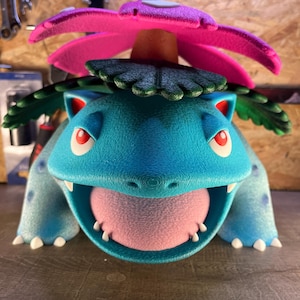 May include: A detailed, three-dimensional sculpture of a Venusaur, a popular character from the Pokémon franchise. The sculpture features a blue body, red eyes, and a large pink flower on its back. The mouth is open, revealing a pink tongue and small white teeth.