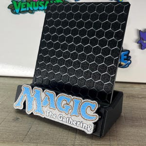 Op de afbeelding: Een zwarte displaystandaard met een gestructureerd zeshoekig rasterpatroon op het achterpaneel. De voorste basis is versierd met een blauw en wit geborduurde patch met de tekst Magic The Gathering in een gestileerd lettertype met een gouden rand.