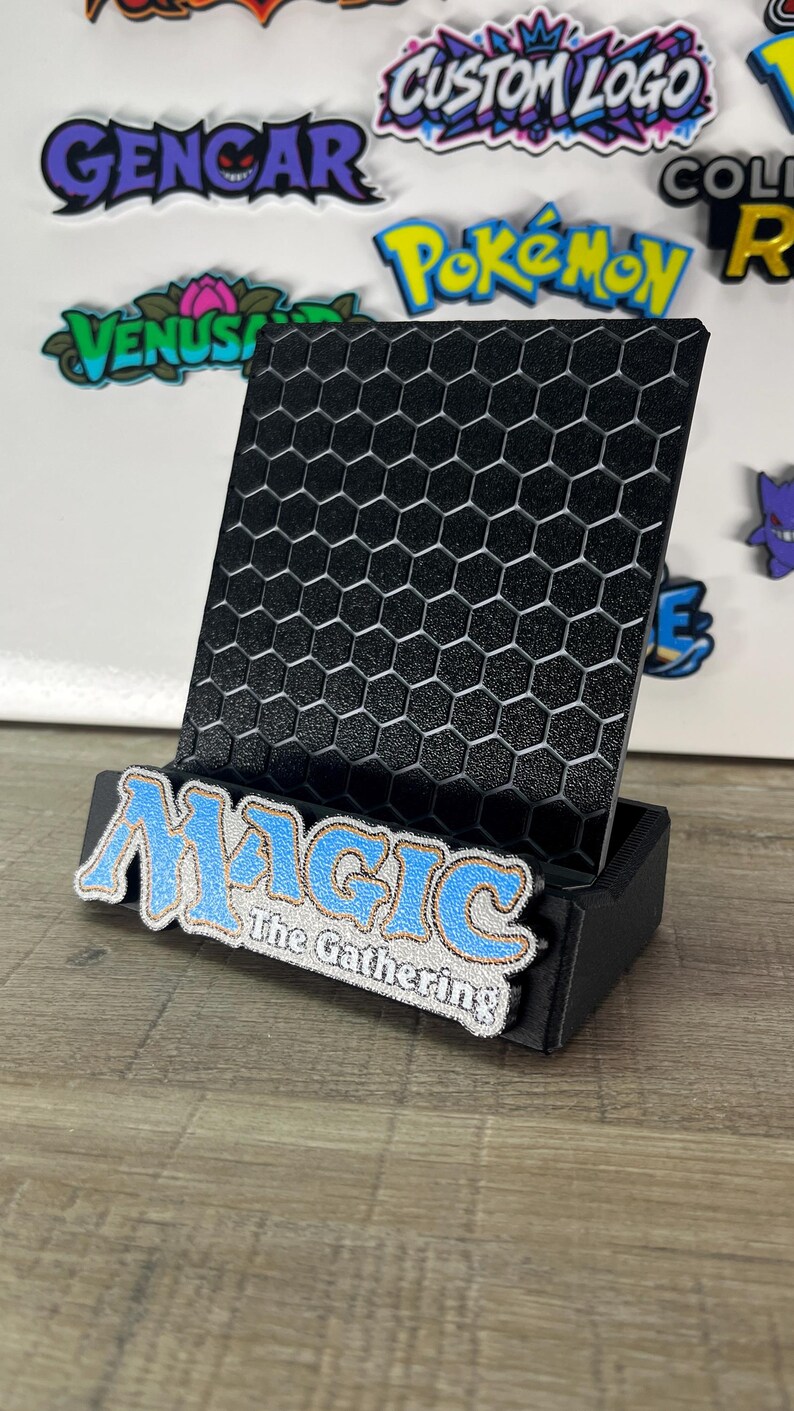 Op de afbeelding: Een zwarte displaystandaard met een gestructureerd zeshoekig rasterpatroon op het achterpaneel. De voorste basis is versierd met een blauw en wit geborduurde patch met de tekst Magic The Gathering in een gestileerd lettertype met een gouden rand.