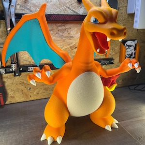 May include: A vibrant orange Charizard figure with blue wings and white claws. The figure has an open mouth revealing a red tongue and teeth. The figure is standing with its arms outstretched, showcasing its detailed design and texture.