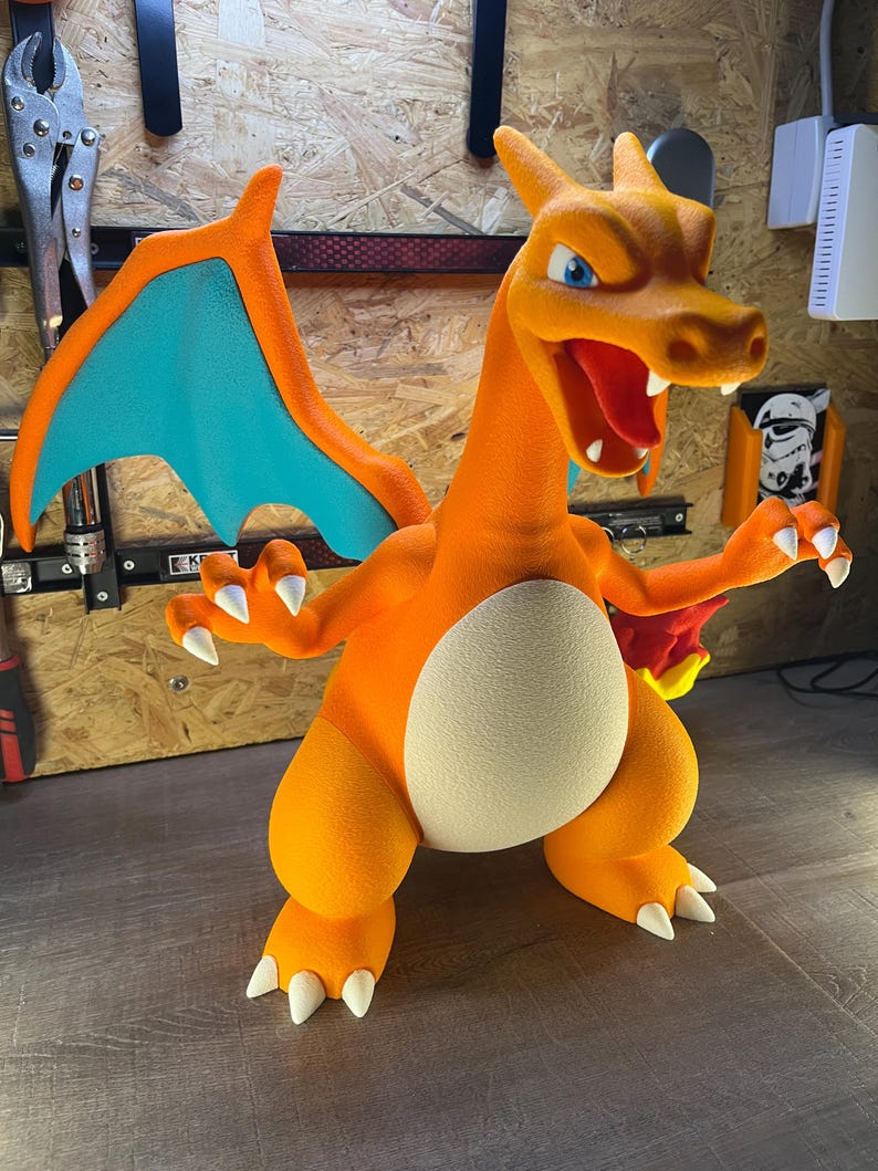 May include: A vibrant orange Charizard figure with blue wings and white claws. The figure has an open mouth revealing a red tongue and teeth. The figure is standing with its arms outstretched, showcasing its detailed design and texture.