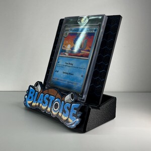 Puede incluir: Un soporte de exhibici&oacute;n negro con patr&oacute;n hexagonal que sostiene una tarjeta coleccionable Wartortle en una funda protectora transparente. El frente tiene un emblema texturizado azul y marr&oacute;n con la palabra BLASTOISE en negrita.