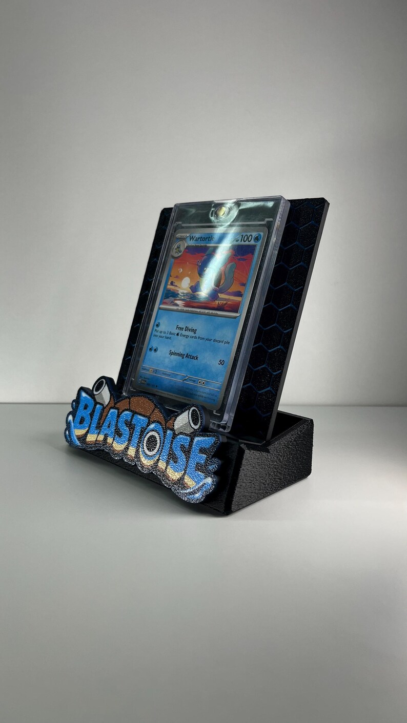 Puede incluir: Un soporte de exhibici&oacute;n negro con patr&oacute;n hexagonal que sostiene una tarjeta coleccionable Wartortle en una funda protectora transparente. El frente tiene un emblema texturizado azul y marr&oacute;n con la palabra BLASTOISE en negrita.