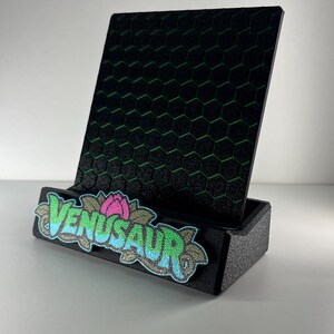 May include: A black display stand featuring a textured hexagonal pattern on the back panel. The front base is adorned with an embroidered patch displaying the text VENUSAUR in blue and green letters, accented by a pink flower and green leaves.