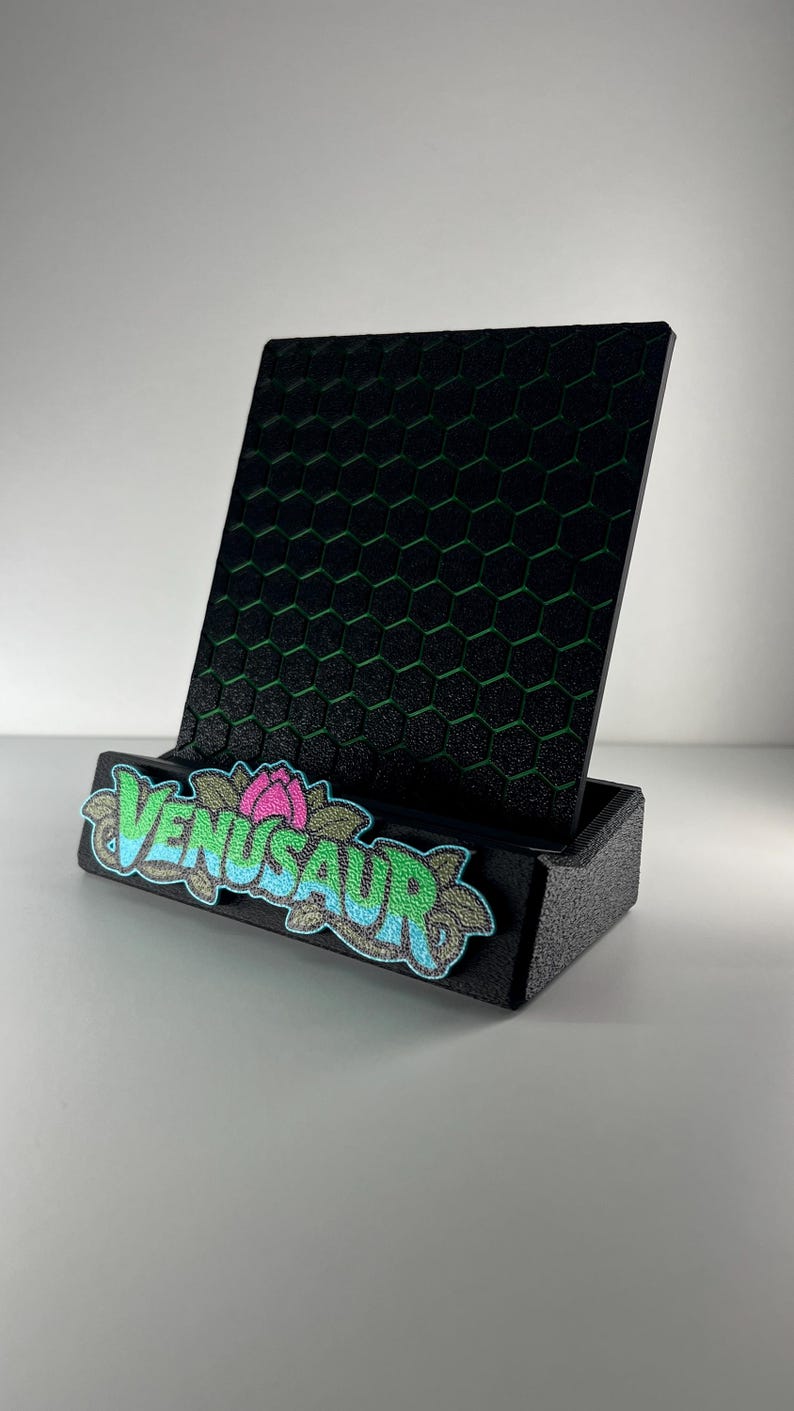 May include: A black display stand featuring a textured hexagonal pattern on the back panel. The front base is adorned with an embroidered patch displaying the text VENUSAUR in blue and green letters, accented by a pink flower and green leaves.