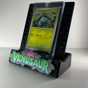May include: A black display stand with a green honeycomb pattern holding a Bisaknosp trading card in a clear protective case. The front of the stand features a textured patch with the word VENUSAUR in blue and green letters.