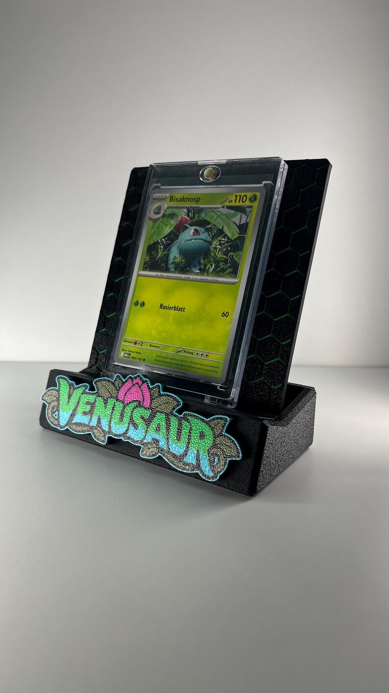 May include: A black display stand with a green honeycomb pattern holding a Bisaknosp trading card in a clear protective case. The front of the stand features a textured patch with the word VENUSAUR in blue and green letters.