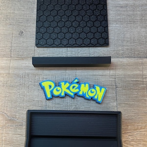 K&ouml;nnte beinhalten: Ein schwarzer quadratischer Pr&auml;sentationssockel mit texturiertem Wabenmuster, ein separater schwarzer rechteckiger St&auml;nder, ein blau-gelbes Pok&eacute;mon-Logo-Emblem und eine schwarze rechteckige Ablage mit gerillter Innenfl&auml;che.