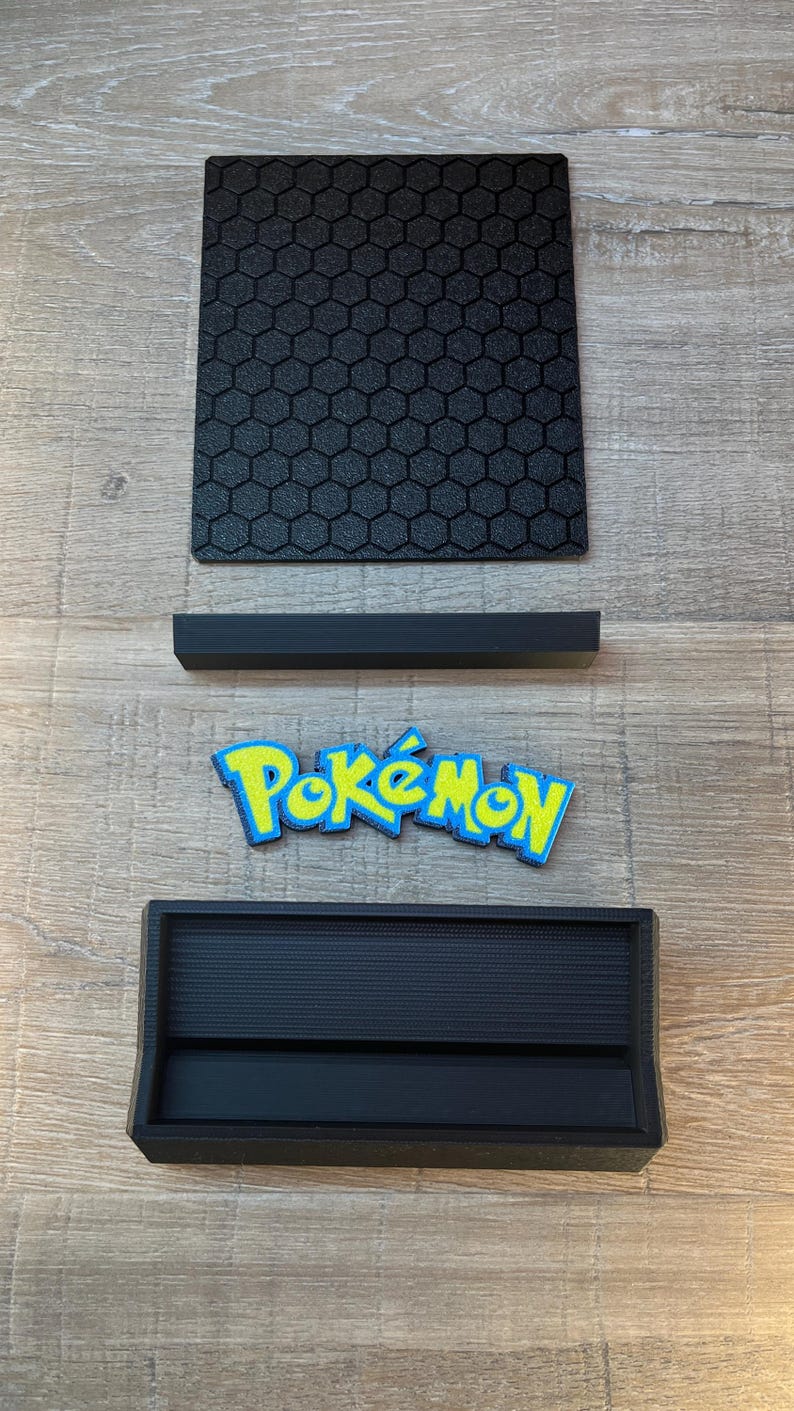 K&ouml;nnte beinhalten: Ein schwarzer quadratischer Pr&auml;sentationssockel mit texturiertem Wabenmuster, ein separater schwarzer rechteckiger St&auml;nder, ein blau-gelbes Pok&eacute;mon-Logo-Emblem und eine schwarze rechteckige Ablage mit gerillter Innenfl&auml;che.
