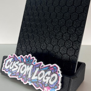 K&ouml;nnte beinhalten: Ein schwarzer Handyhalter mit einem strukturierten Sechseckmuster auf der R&uuml;ckseite. An der Vorderseite ist ein wei&szlig;es, ausgestanztes Schild mit dem schwarzen Schriftzug "CUSTOM LOGO" und einem bunten Graffiti-Design in Blau, Pink und Lila angebracht.
