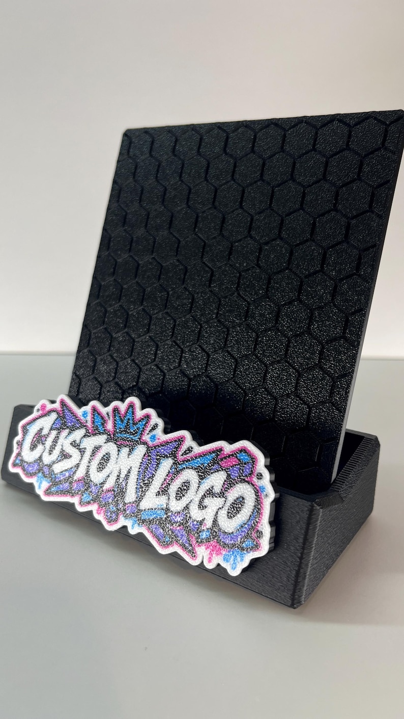 K&ouml;nnte beinhalten: Ein schwarzer Handyhalter mit einem strukturierten Sechseckmuster auf der R&uuml;ckseite. An der Vorderseite ist ein wei&szlig;es, ausgestanztes Schild mit dem schwarzen Schriftzug "CUSTOM LOGO" und einem bunten Graffiti-Design in Blau, Pink und Lila angebracht.