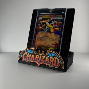 May include: A black display stand featuring a prominent Charizard logo with blue and orange wings. The stand holds a Mega Charizard Y EX trading card encased in a clear protective magnetic holder, set against a black hexagonal patterned backplate.
