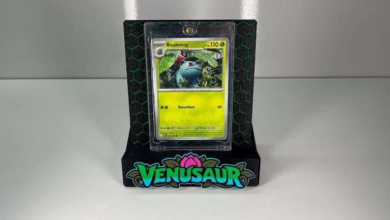 May include: A Bisaknosp trading card encased in a clear protective magnetic holder, mounted on a black display stand with a green hexagonal pattern and the word VENUSAUR in large green and blue lettering at the base.