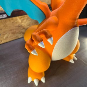 May include: A close-up of an orange and white figurine of a Charizard. The figurine has white claws, a white belly, and blue wings. The figurine is on a gray surface.