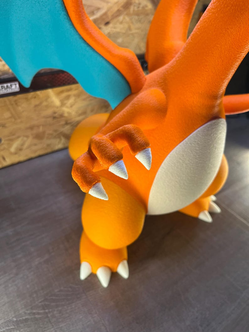 May include: A close-up of an orange and white figurine of a Charizard. The figurine has white claws, a white belly, and blue wings. The figurine is on a gray surface.