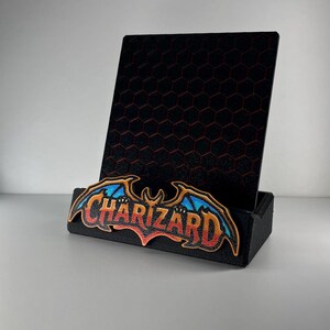 May include: A black display stand featuring a vertical back panel with a red hexagonal grid pattern. The front base is adorned with a colorful embroidered patch displaying the word CHARIZARD in stylized orange, red, and blue lettering.