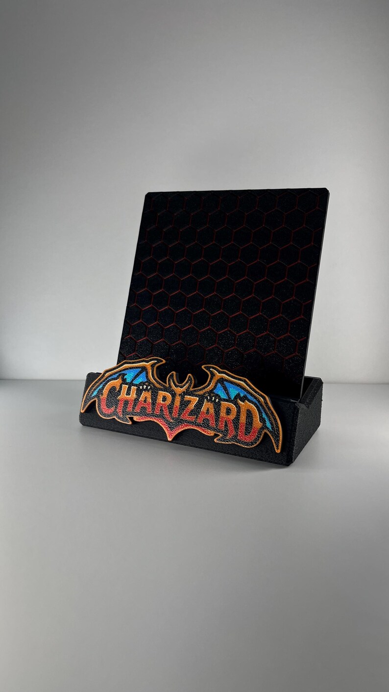 May include: A black display stand featuring a vertical back panel with a red hexagonal grid pattern. The front base is adorned with a colorful embroidered patch displaying the word CHARIZARD in stylized orange, red, and blue lettering.