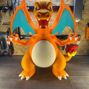 May include: A large, orange Charizard figure with blue wings and white claws. The figure has a cream-colored belly and a flame on its tail. The figure is posed with its arms outstretched and mouth open.