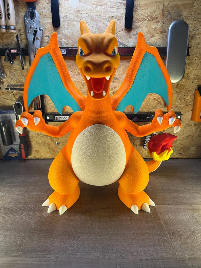 May include: A large, orange Charizard figure with blue wings and white claws. The figure has a cream-colored belly and a flame on its tail. The figure is posed with its arms outstretched and mouth open.