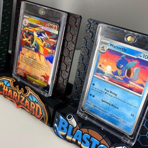 May include: Three black display stands for trading cards, each featuring a hexagonal pattern and a colorful nameplate reading Venusaur, Charizard, or Blastoise. Each stand holds a clear protective case containing a specific Pok&eacute;mon card.
