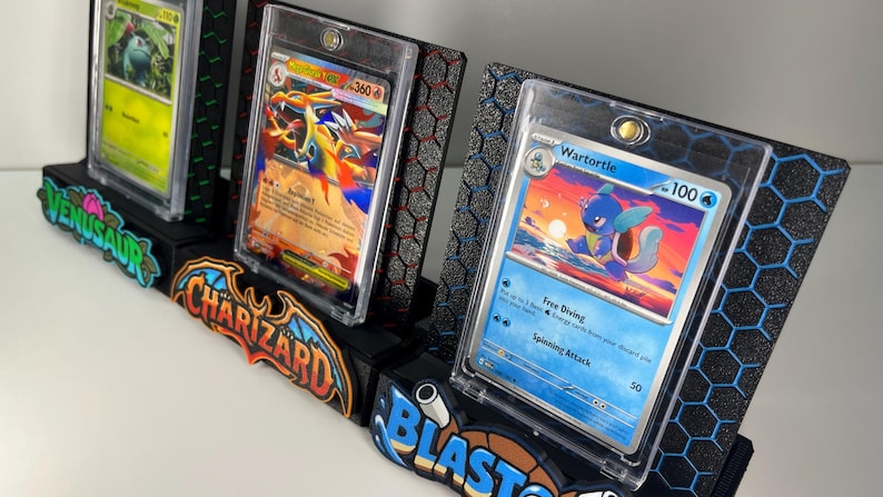 May include: Three black display stands for trading cards, each featuring a hexagonal pattern and a colorful nameplate reading Venusaur, Charizard, or Blastoise. Each stand holds a clear protective case containing a specific Pok&eacute;mon card.