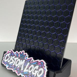 K&ouml;nnte beinhalten: Ein schwarzer Handyhalter mit texturiertem Wabenmuster und violetten Akzenten. Die vordere Basis zeigt eine wei&szlig;e, blaue und violette Grafikplatte mit dem Text CUSTOM LOGO in einer stilisierten, glitzernden Graffiti-Schriftart.