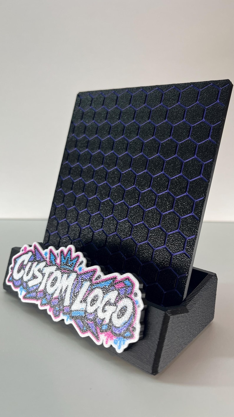 K&ouml;nnte beinhalten: Ein schwarzer Handyhalter mit texturiertem Wabenmuster und violetten Akzenten. Die vordere Basis zeigt eine wei&szlig;e, blaue und violette Grafikplatte mit dem Text CUSTOM LOGO in einer stilisierten, glitzernden Graffiti-Schriftart.
