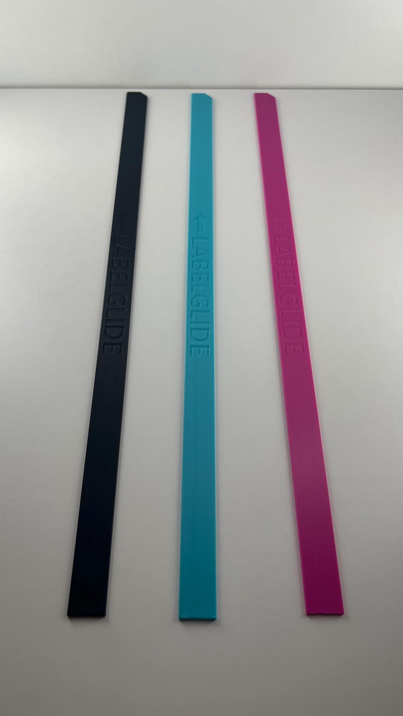 Peut inclure: Trois longues bandes rectangulaires et fines de couleur noire, bleu vif et magenta. Chaque bande pr&eacute;sente le texte en relief LABELGLIDE orient&eacute; verticalement sur toute la longueur, accompagn&eacute; d'une petite ic&ocirc;ne de fl&egrave;che pointant vers le texte.