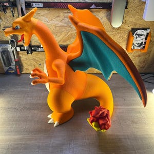 May include: A large, orange Charizard figure with teal wings and white claws. The dragon figure has a long neck, a detailed face, and a flame at the end of its tail. The figure is a collectible item.