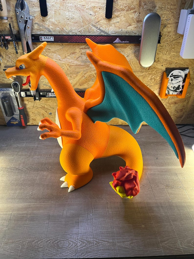 May include: A large, orange Charizard figure with teal wings and white claws. The dragon figure has a long neck, a detailed face, and a flame at the end of its tail. The figure is a collectible item.