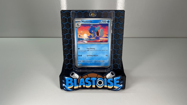 Puede incluir: Una carta coleccionable de Pokemon Wartortle en una funda protectora transparente, montada en un soporte negro con el logotipo de Blastoise en azul y beige en la base. El soporte presenta un patr&oacute;n de panal negro en el panel trasero.