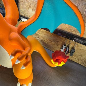 May include: Orange and blue Charizard figure with white claws and a flame-shaped tail. The wings are orange with blue undersides. Two keychains with the word "MOM" are visible.