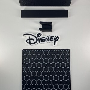 May include: A black rectangular tray, a thin black rectangular strip, a small black U-shaped bracket, a white and black Disney logo cutout, and a black square base featuring a raised gray honeycomb pattern on the surface.
