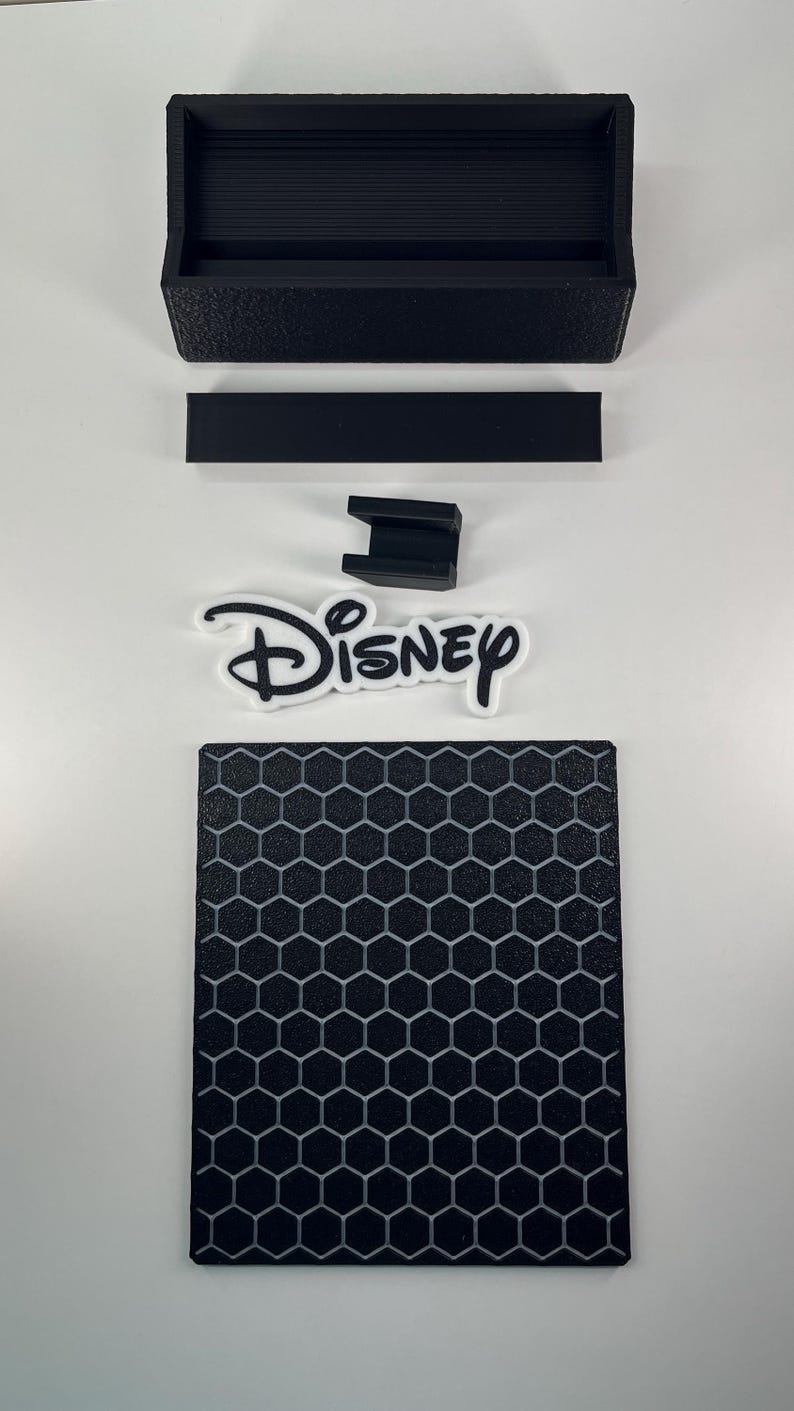 May include: A black rectangular tray, a thin black rectangular strip, a small black U-shaped bracket, a white and black Disney logo cutout, and a black square base featuring a raised gray honeycomb pattern on the surface.