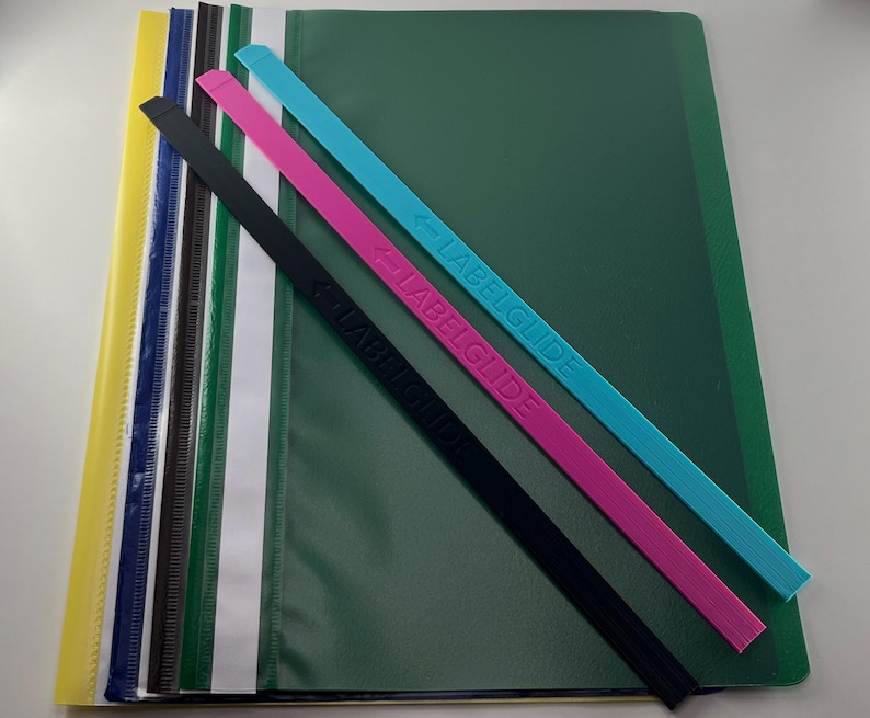 Peut inclure: Une collection de chemises &agrave; documents en plastique multicolores avec des bandes en plastique fines et allong&eacute;es assorties portant l'inscription LABELGUIDE. Les chemises sont empil&eacute;es en jaune, bleu, noir, blanc et vert, avec des bandes diagonales.